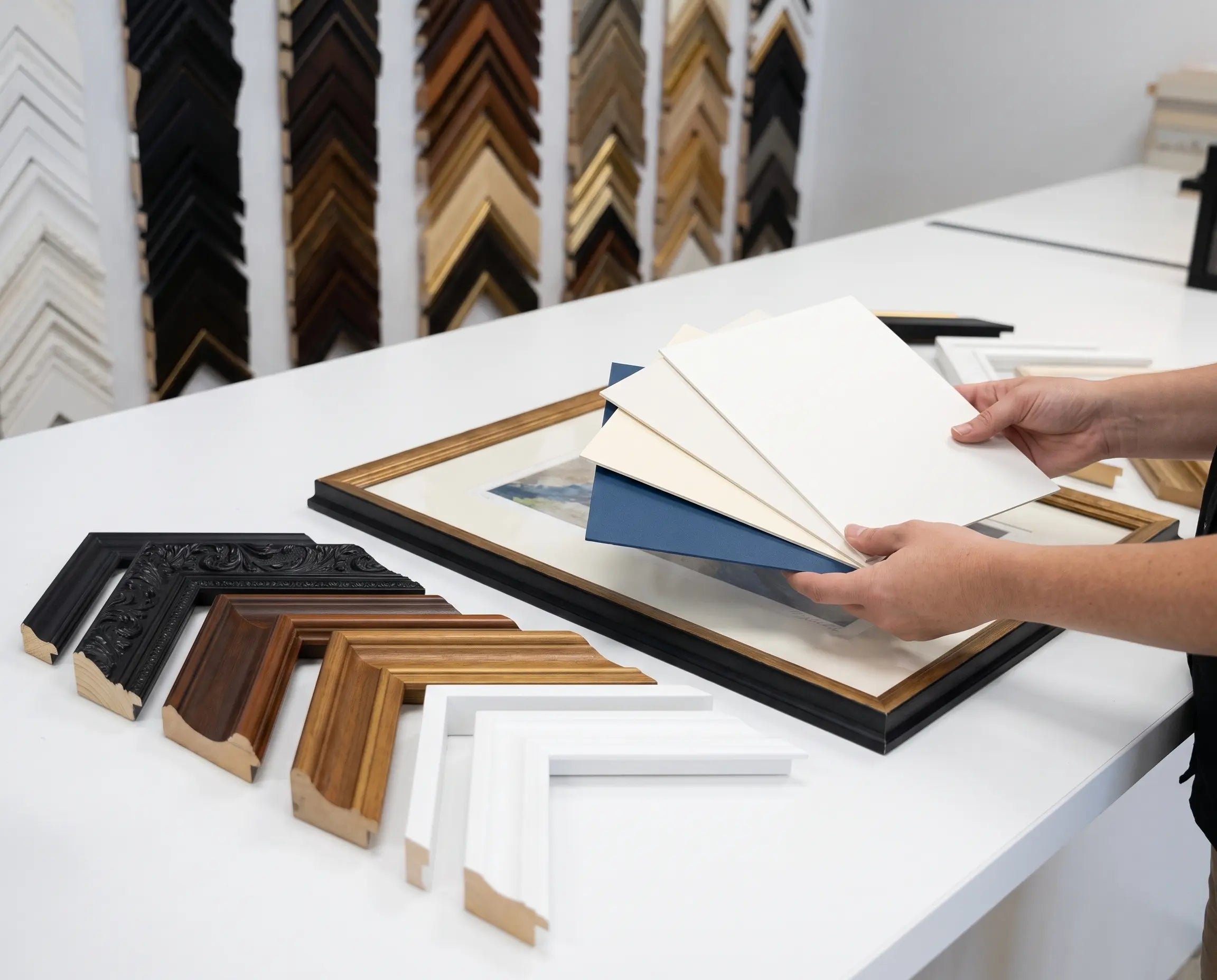 A person's hands holding a stack of white and blue sample boards in front of a picture frame on a white table, with various picture frame samples displayed on stands in the background.