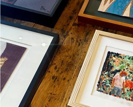 Several framed artworks arranged on a rustic wooden table. The pieces include colorful abstract and figurative paintings in different frame styles: black frames, light wood frames, and white mats.